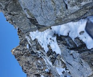 Snowy mountain path with rocky terrain and a safety chain along the side under a clear blue sky.