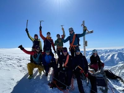 Gruppe von Bergsteigern mit Helmen und Seilen auf schneebedecktem Gipfel mit Bergpanorama im Hintergrund.