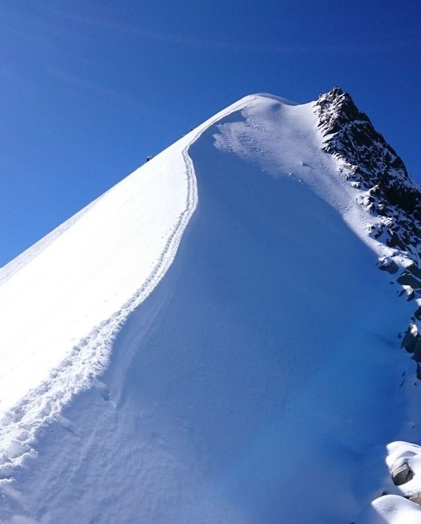 Snow-covered mountain peak with a narrow ridge path under a clear blue sky.