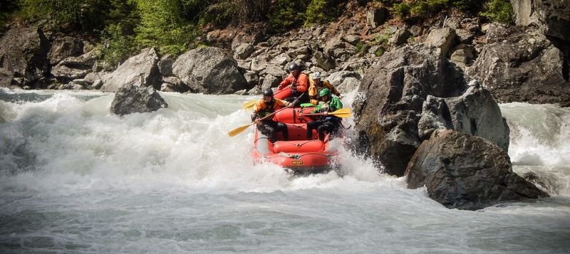 Personen in einem Schlauchboot beim Wildwasser-Rafting, tragen Helme und Schwimmwesten, Felsen.