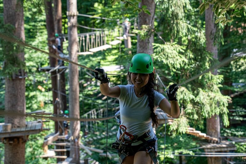 Person wearing a green helmet and harness navigating a rope course in a dense forest adventure park.
