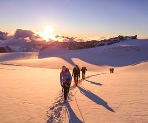Hikers with helmets and harnesses trek across a snowy mountain landscape at sunrise.