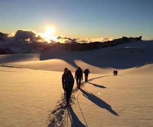 Hikers with helmets and harnesses walk on a snowy mountain path at sunrise, casting long shadows.