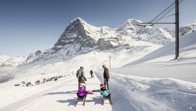 People are sledding in the snow in front of a mountain backdrop, wearing winter clothing and helmets.