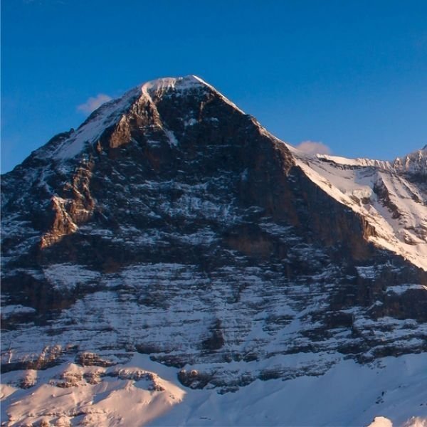 A group of people with backpacks and poles hike up a snowy mountain slope near a rocky peak.