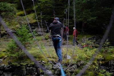 Person on rope bridge in the forest with climbing harness and helmet; two people are standing on the ground.