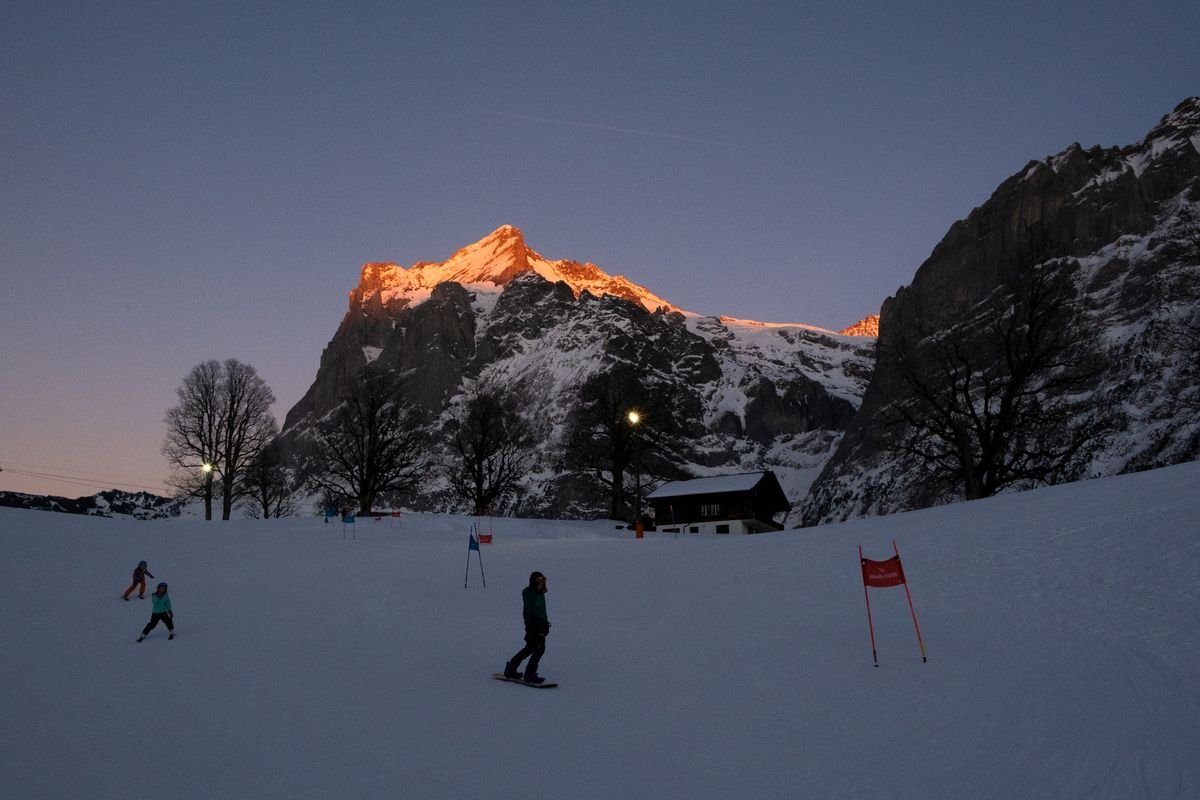 People are skiing on a slope at sunset, surrounded by snow-covered mountains.