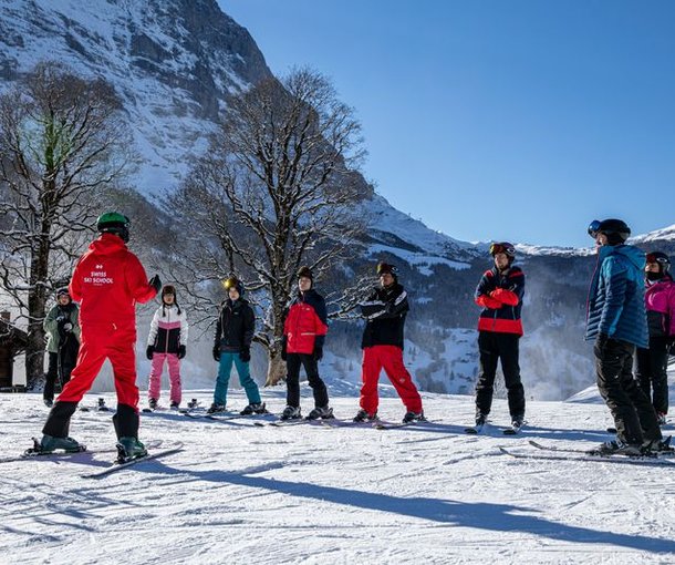 Ein Skilehrer in einem roten Anzug führt eine Gruppe von Skifahrern mit Helmen und Skibrillen auf einem verschneiten Berghang.