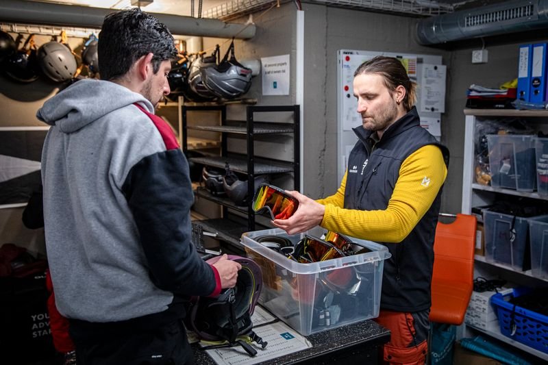 Zwei Personen in einem Ausrüstraum, eine Person reicht der anderen Skibrillen, umgeben von Helmen.