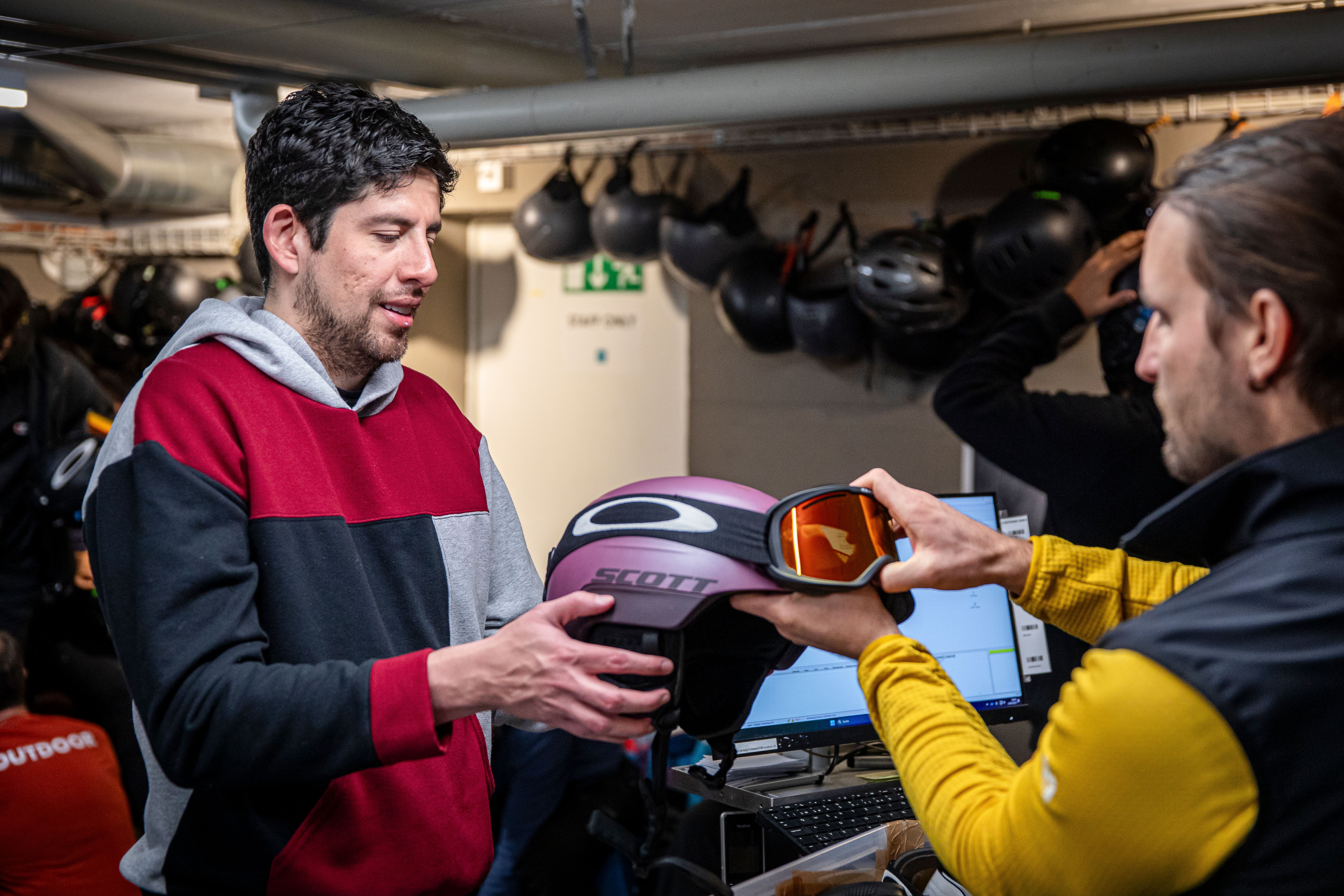 Zwei Personen tauschen einen Skihelm mit Skibrille in einem Innenbereich zur Ausrüstungsvermietung mit Helmen auf Regalen aus.