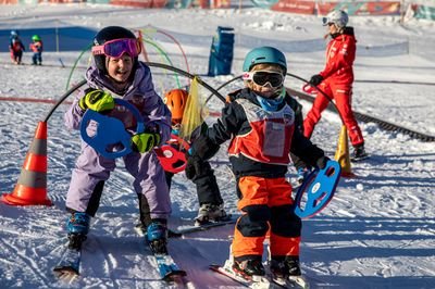 Two children with helmets and skis on a slope, accompanied by a ski instructor in red clothing.