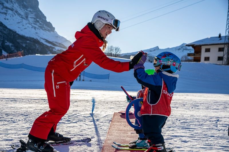 Skilehrer in roter Ausrüstung gibt Kind mit Helm und Skiern auf schneebedecktem Hang Anweisungen.