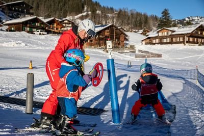 Two children with helmets and skis on a conveyor belt, accompanied by an adult in red ski clothing.