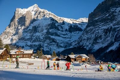 Children with skis and helmets on a ski slope, accompanied by an adult, in front of snow-covered mountains.