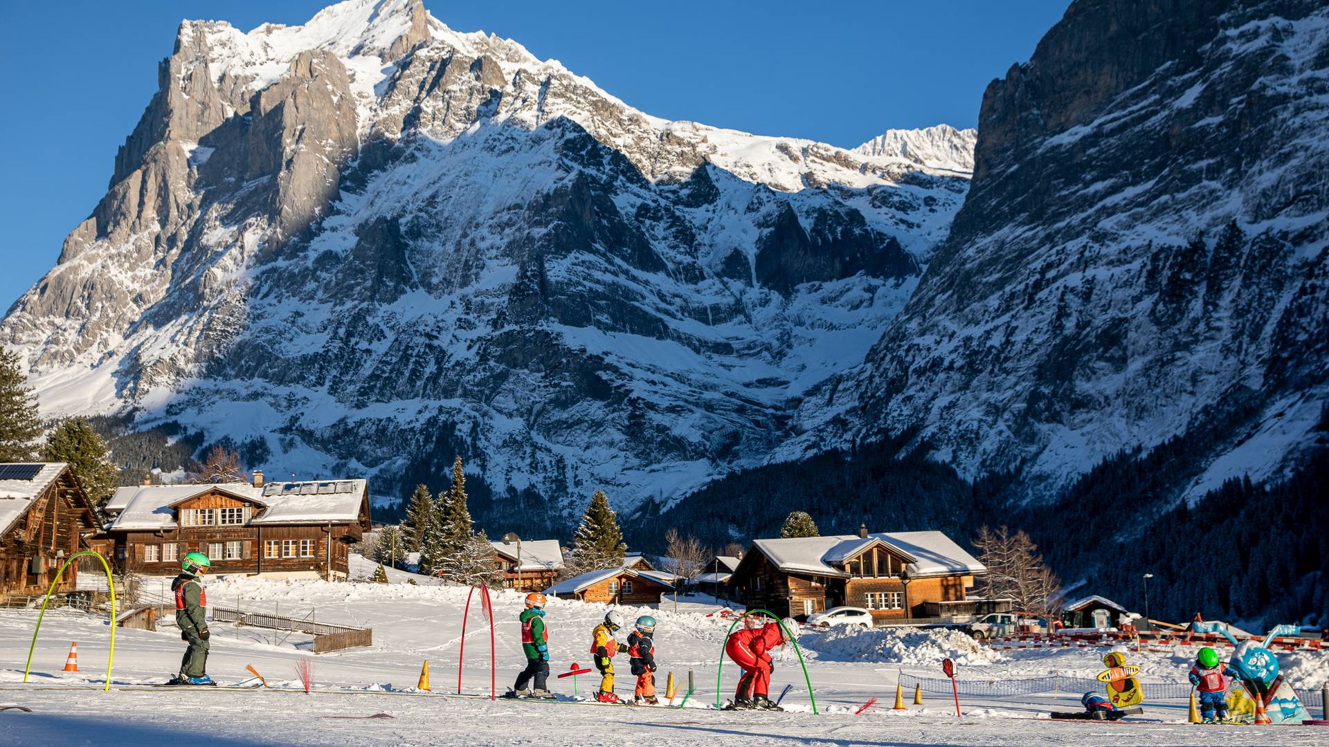 Kinder mit Skiern und Helmen auf Skipiste, begleitet von einem Erwachsenen, vor schneebedeckten Bergen.