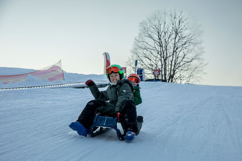 Two people are sledding on a snow-covered slope, wearing helmets and winter clothing.