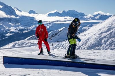 People with skis on a slope in the mountains; one person is wearing red clothing, the other black.