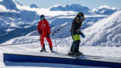 People with skis on a slope in the mountains; one person is wearing red clothing, the other black.