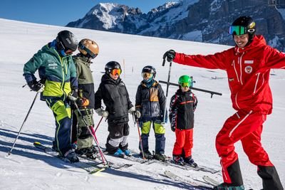 Ski group with helmets and ski poles on a snowy mountain, accompanied by a ski instructor in red.