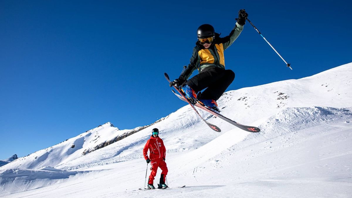 People skiing on a snow-covered mountain; one person is jumping, both wearing helmets and skis.