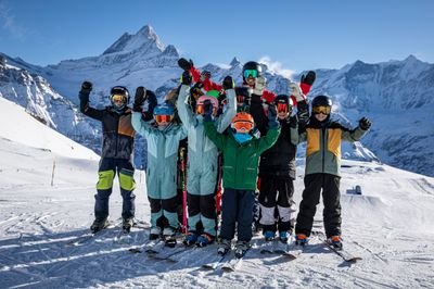 Gruppe von Skifahrern mit Helmen und Skibrillen auf schneebedecktem Berg vor alpiner Kulisse.