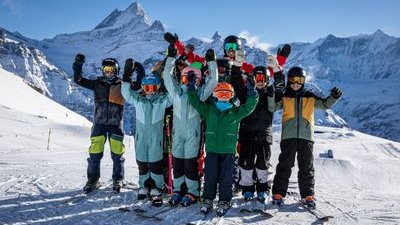 Gruppe von Skifahrern mit Helmen und Skibrillen auf schneebedecktem Berg vor alpiner Kulisse.