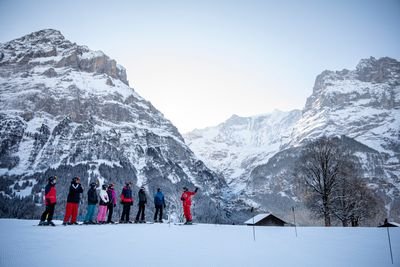 Skiers in helmets and gear stand on a snowy slope with a guide, surrounded by snowy mountains.