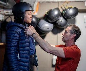 Person in Helm und Jacke wird von einer anderen Person in einem Raum mit Helmen an der Wand unterstützt.