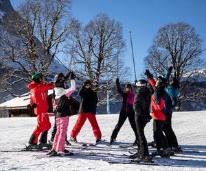 Skifahrer in Helmen und Ausrüstung heben Stöcke in einer schneebedeckten Berglandschaft mit Bäumen und einem Chalet.