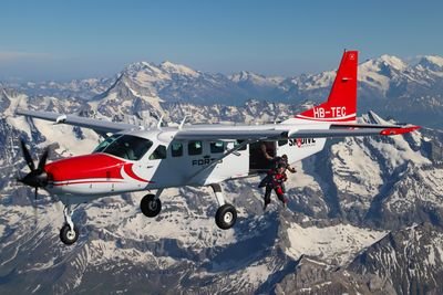 Fallschirmspringer mit Helm und Anzug springt aus Flugzeug über schneebedeckten Alpen.