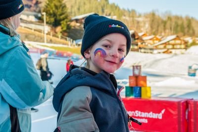 Child on ski slope with painted face, beanie, and winter jacket; snow-covered mountains in the background.