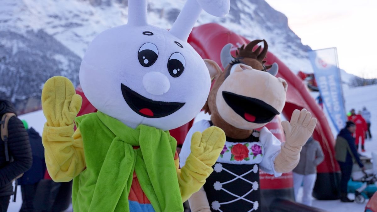 Zwei Maskottchen in bunten Kostümen auf einer schneebedeckten Berglandschaft, Menschen im Hintergrund.