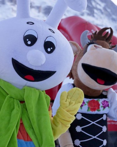 Zwei Maskottchen in bunten Kostümen auf einer schneebedeckten Berglandschaft, Menschen im Hintergrund.