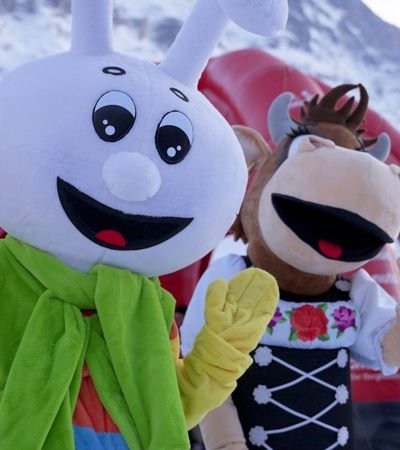 Two mascots in colorful costumes on a snow-covered mountain landscape, people in the background.