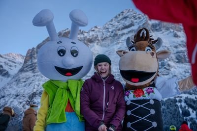 Person in winter clothing with two mascots in front of snow-covered mountains.