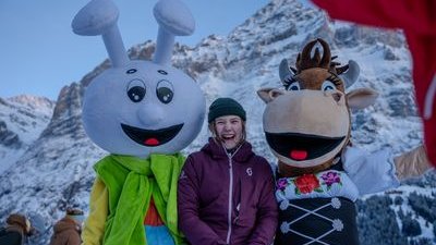 Person in winter clothing with two mascots in front of snow-covered mountains.