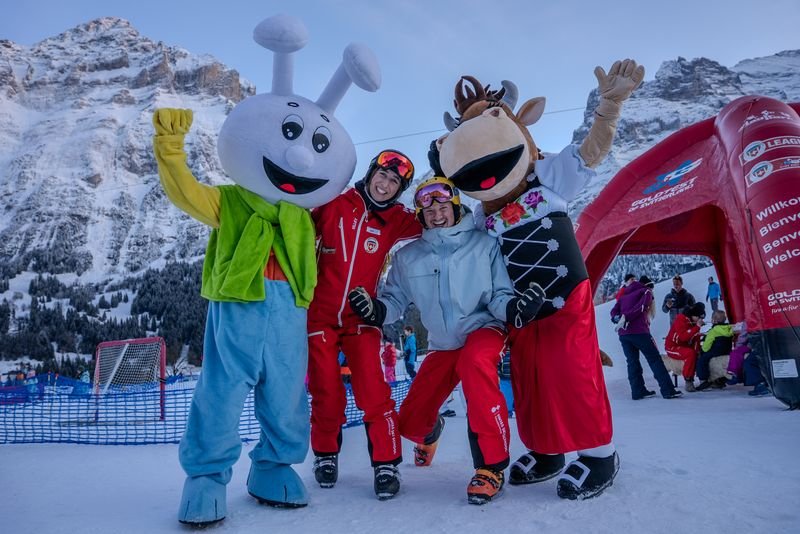 People in ski gear posing with two mascots in front of a mountain backdrop in the snow.