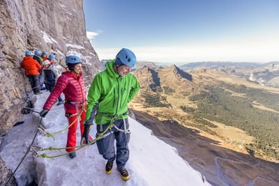People with helmets and climbing harnesses on a snowy mountain path, secured with ropes.