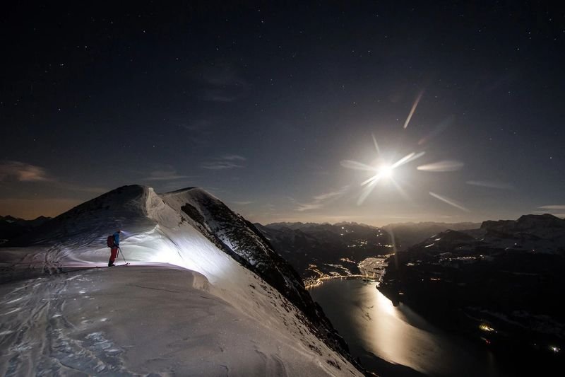 Skier with poles and backpack on snowy mountain ridge at night, overlooking a lake and distant lights.