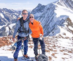 Two people in climbing gear stand on a snowy mountain peak with trekking poles and backpacks.