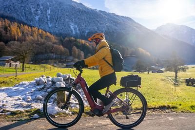 Person cycling on a paved path, wearing a helmet and backpack, with snowy mountains in the background.