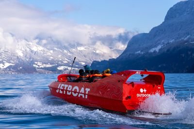 Red jet ski with people in life jackets on a lake, surrounded by snow-covered mountains.