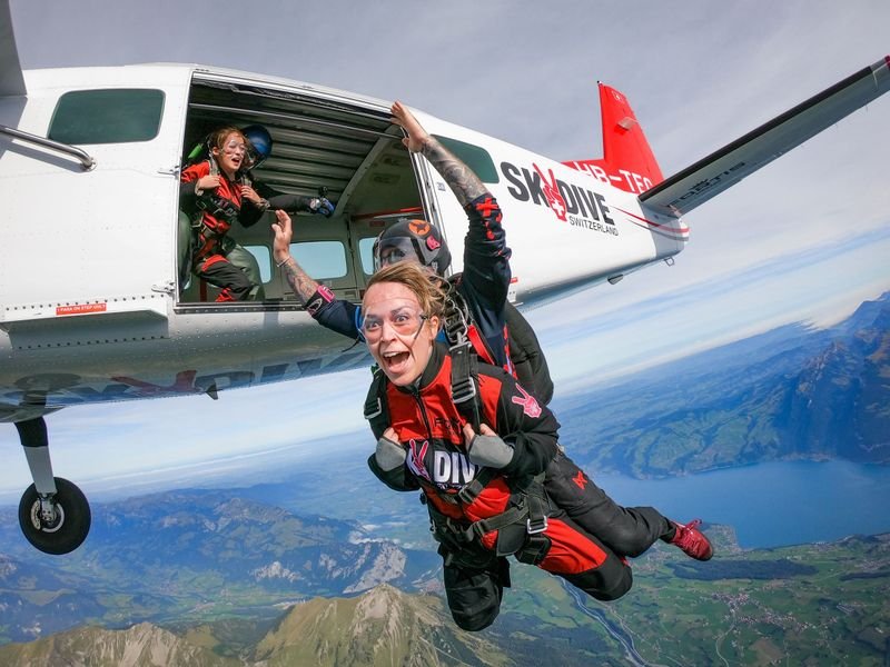 Tandem skydivers in red jumpsuits and goggles exit a plane over mountains and a lake in Switzerland.