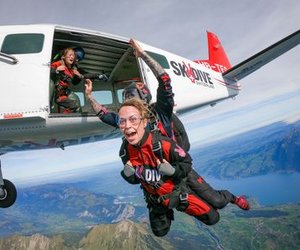 Fallschirmspringer im Tandem in roten Sprunganzügen und Schutzbrillen verlassen ein Flugzeug über Bergen und einem See in der Schweiz.