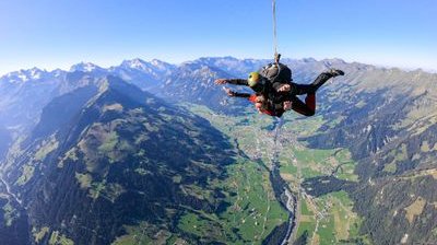 Zwei Personen machen Tandem-Fallschirmspringen über einer bergigen Landschaft, tragen Helme und Gurte.
