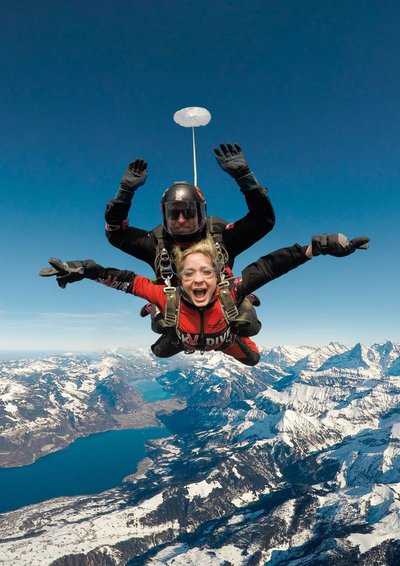 Zwei Personen machen Tandem-Fallschirmspringen über schneebedeckten Bergen, tragen Helme und Gurte.