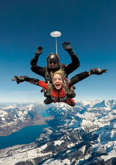 Zwei Personen machen Tandem-Fallschirmspringen über schneebedeckten Bergen, tragen Helme und Gurte.