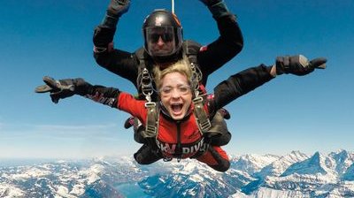 Zwei Personen machen Tandem-Fallschirmspringen über schneebedeckten Bergen, tragen Helme und Gurte.