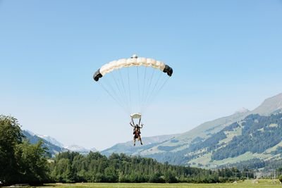 Zwei Personen gleiten mit einem Gleitschirm über einem grasbewachsenen Feld, im Hintergrund Berge.