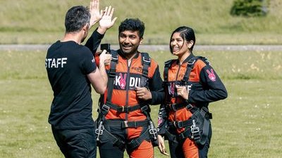 Zwei Personen in Fallschirmspringeranzügen und Gurten stehen auf Gras und interagieren mit einem Mitarbeiter.